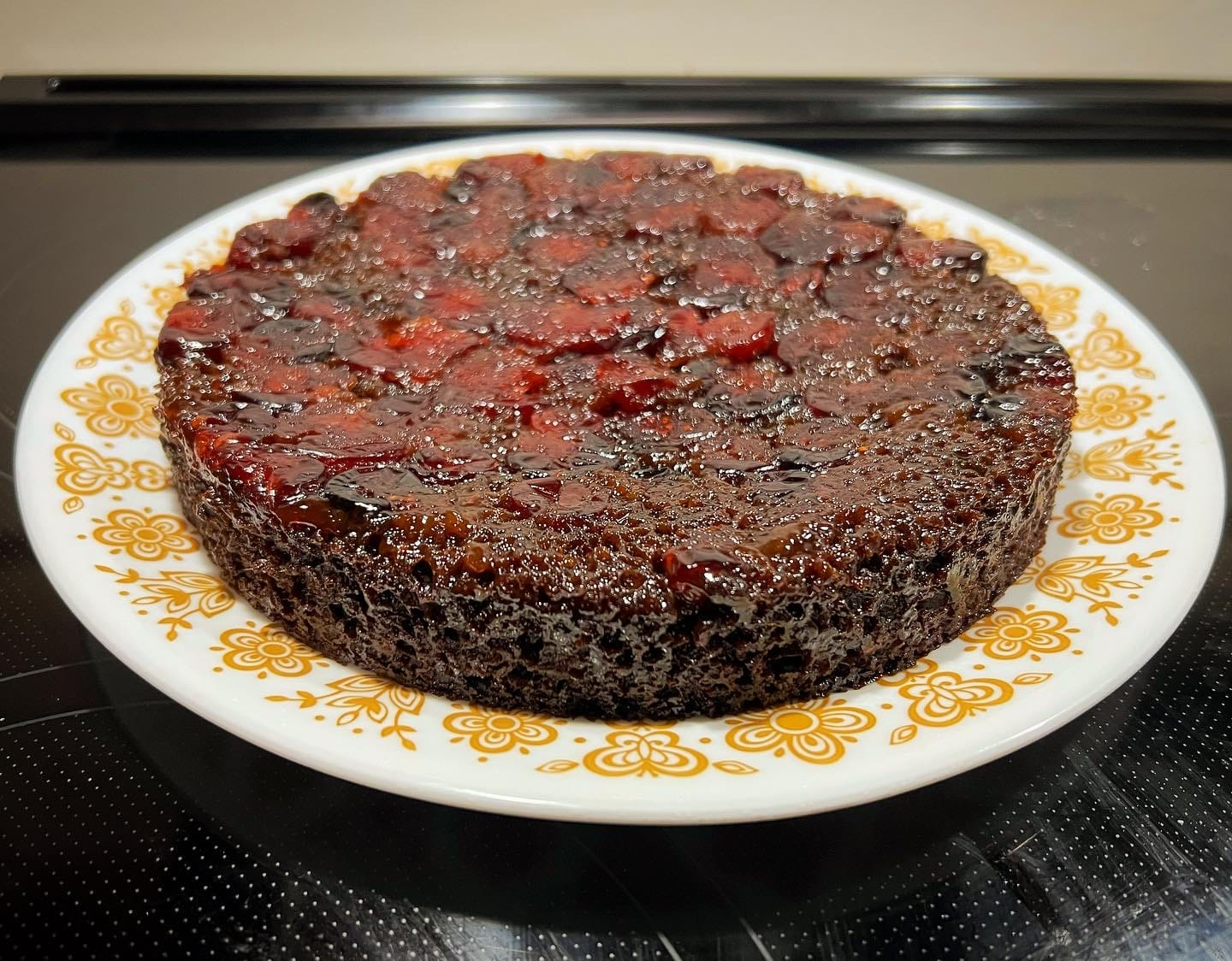 A low, wide cake on a plate. The cake is dark brown and glossy, studded with red cranberries on top.
