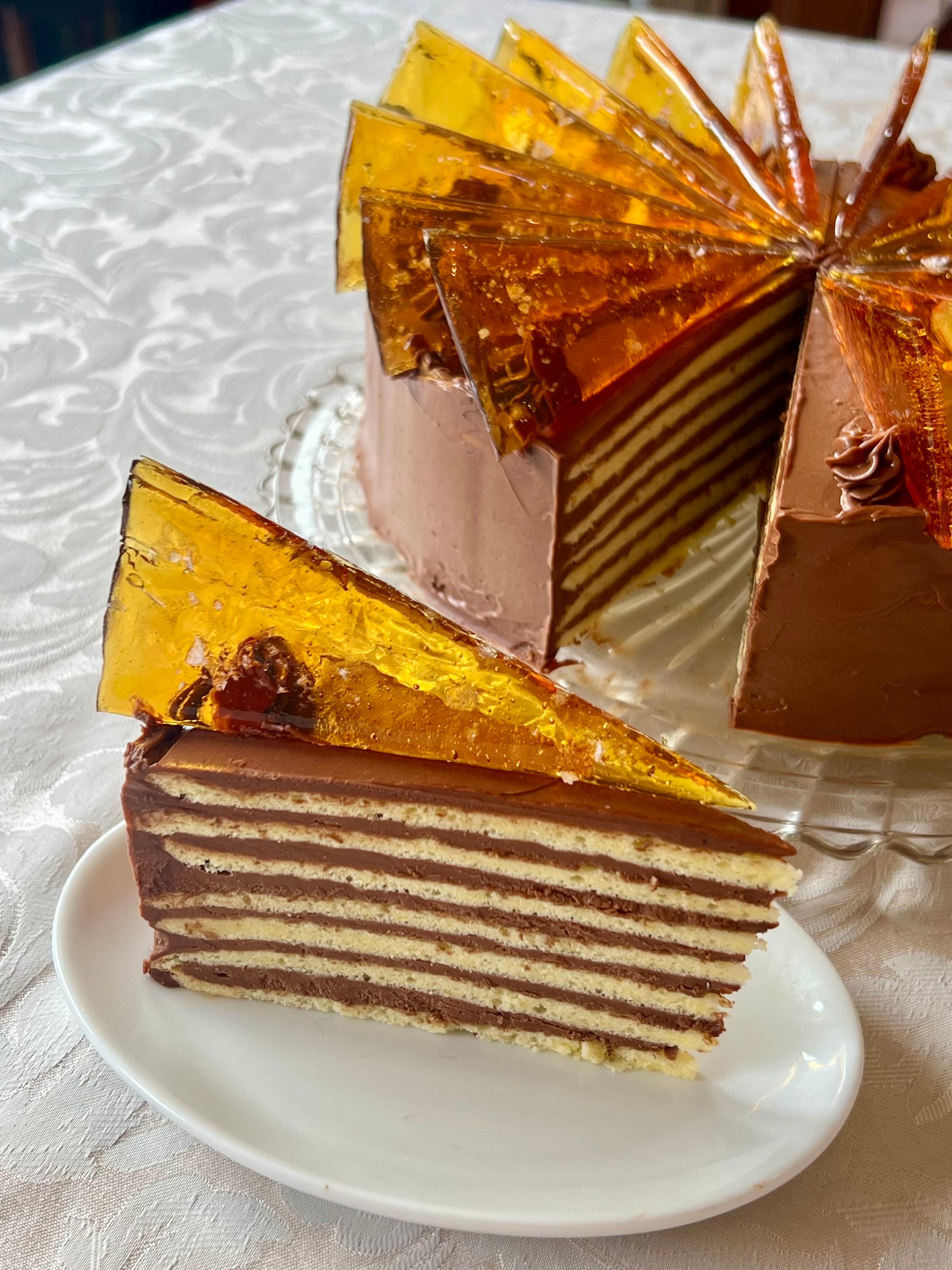 A seven-layer slice of Dobos torta, topped with a triangle of hard caramel, in front of the full cake.