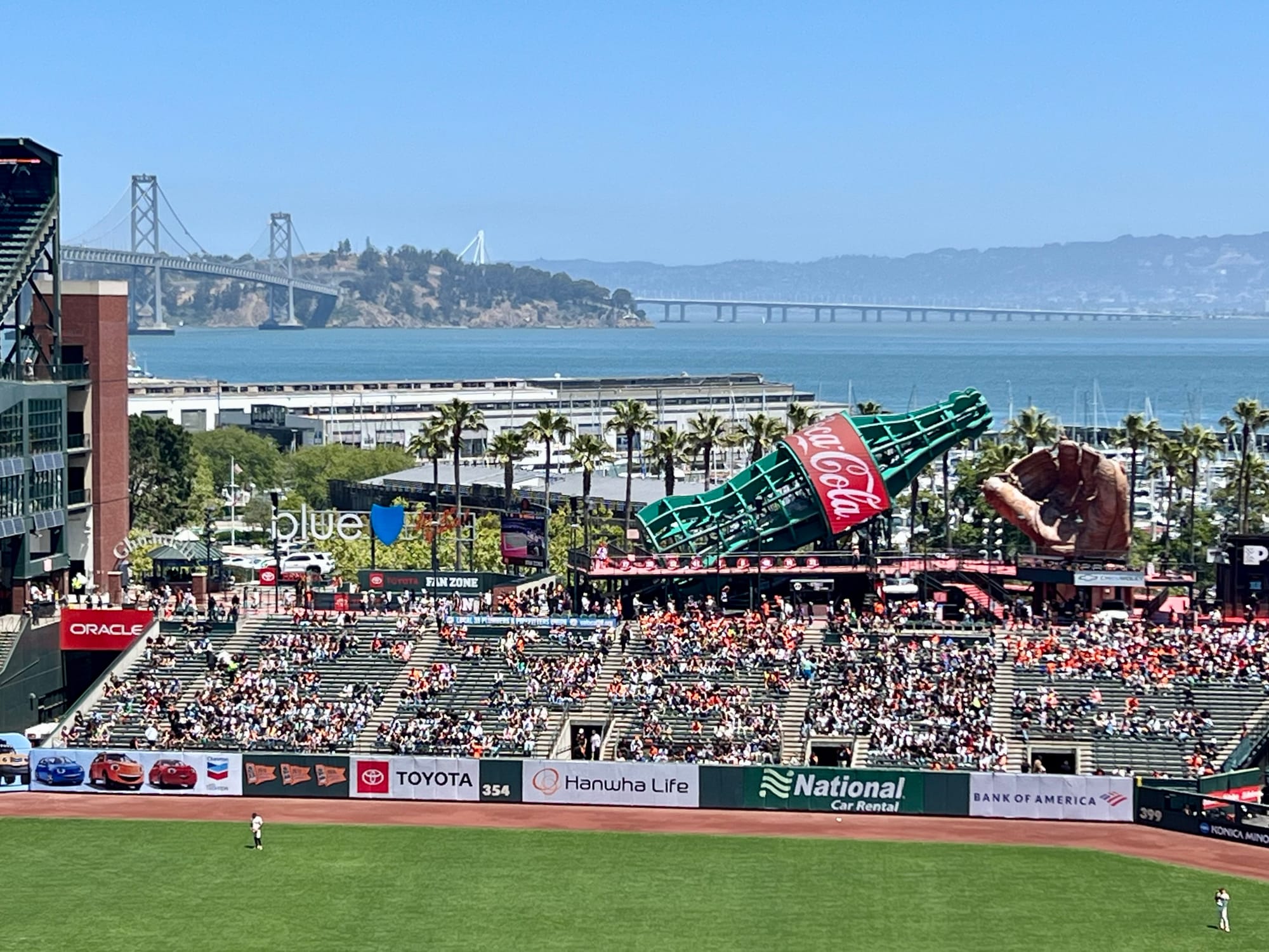 View of one edge of Oracle Park, with giant sculptures of a Coke bottle and a baseball mitt. The Bay Bridge is seen in the distance.
