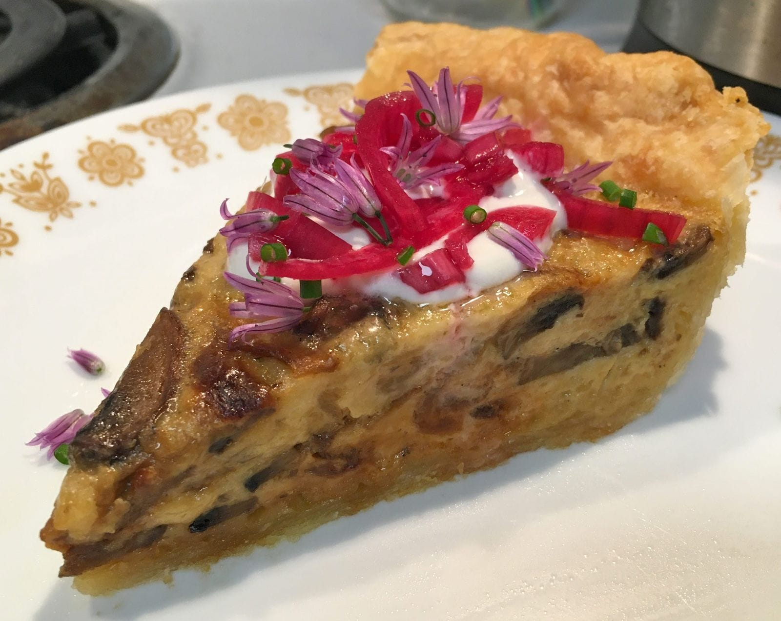 A slice of mushroom and gruyere quiche, topped with creme fraiche, pickled red onions, and chive blossoms.