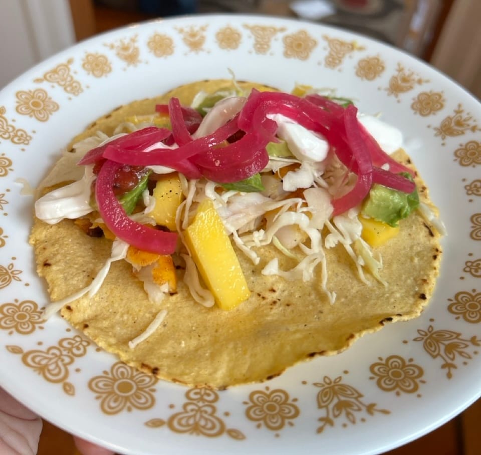 A small fish taco on a corn tortilla, with mango, shredded cabbage, and pickled red onions.