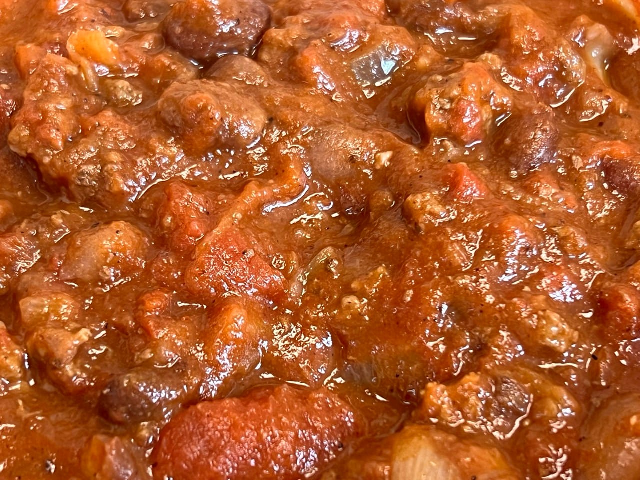 Close-up of the chili. You can see tomatoes, bits of ground beef, beans, and onions, all in a thick, reddish-brown liquid.